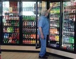 Older man arranging products in an open multi-glass door refrigerator at a convenience store, facing the neatly displayed items.